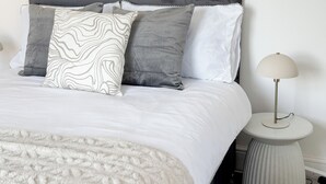 Un lit bien fait, avec une tête de lit grise, des oreillers blancs et gris, et un couvre-lit blanc. Une petite table ronde avec une lampe se trouve à côté du lit.