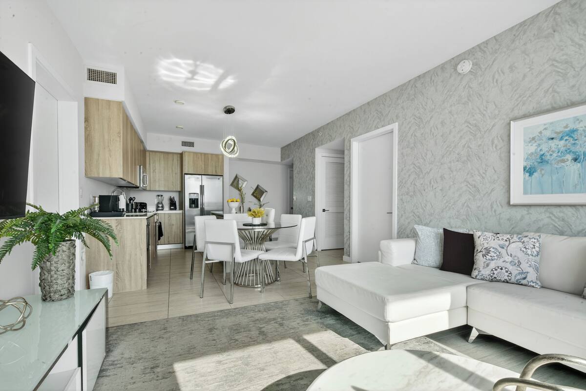 A modern living room with a white sofa, a dining area with a glass table, and a kitchen with wooden cabinets.
