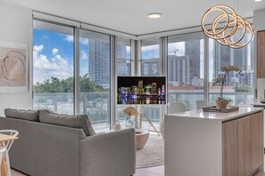 A modern living room with a large TV, a grey sofa, a wooden cabinet, and a view of the city skyline through large windows. - Luxe Condos - Miami Design District (Miami)