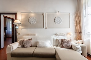 A modern living room with a beige sofa, decorative pillows, wall art, and a side table with a lamp and a welcome sign.