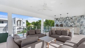 A modern living room with a sofa set, a coffee table, and a dining area with chairs. There is a ceiling fan, large windows, and a view of a pool and buildings outside. - Oceanfront Sunset Patio w/ BBQ + Pool, Gym & Spa (West Bay)