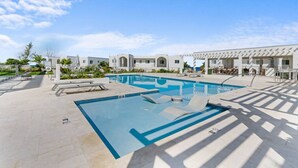 A swimming pool with lounge chairs, a pergola, and a building with a white facade. - Oceanfront Sunset Patio w/ BBQ + Pool, Gym & Spa (West Bay)