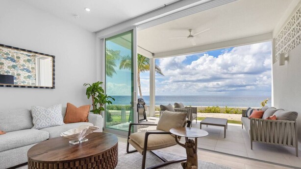 A modern living room with a large glass door leading to a balcony overlooking the ocean. - Oceanfront Sunset Patio w/ BBQ + Pool, Gym & Spa (West Bay)