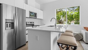 A modern kitchen with a central island, stainless steel appliances, and a large window with a view of greenery. - Oceanfront Sunset Patio w/ BBQ + Pool, Gym & Spa (West Bay)