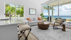 A modern living room with a kitchen area, large windows, and a view of the ocean. - Oceanfront Sunset Patio w/ BBQ + Pool, Gym & Spa (West Bay)