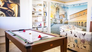 A pool table with a red puck, a sofa with a floral pattern, and a bookshelf with various items.