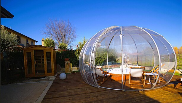 A transparent dome structure with seating inside, a wooden deck, and a small shed in the background.
