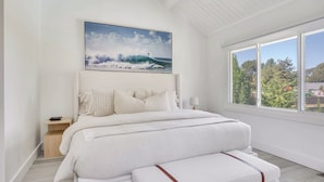 Une chambre avec un grand lit, une table de chevet, une fenêtre donnant sur des arbres et un tableau représentant une vague.