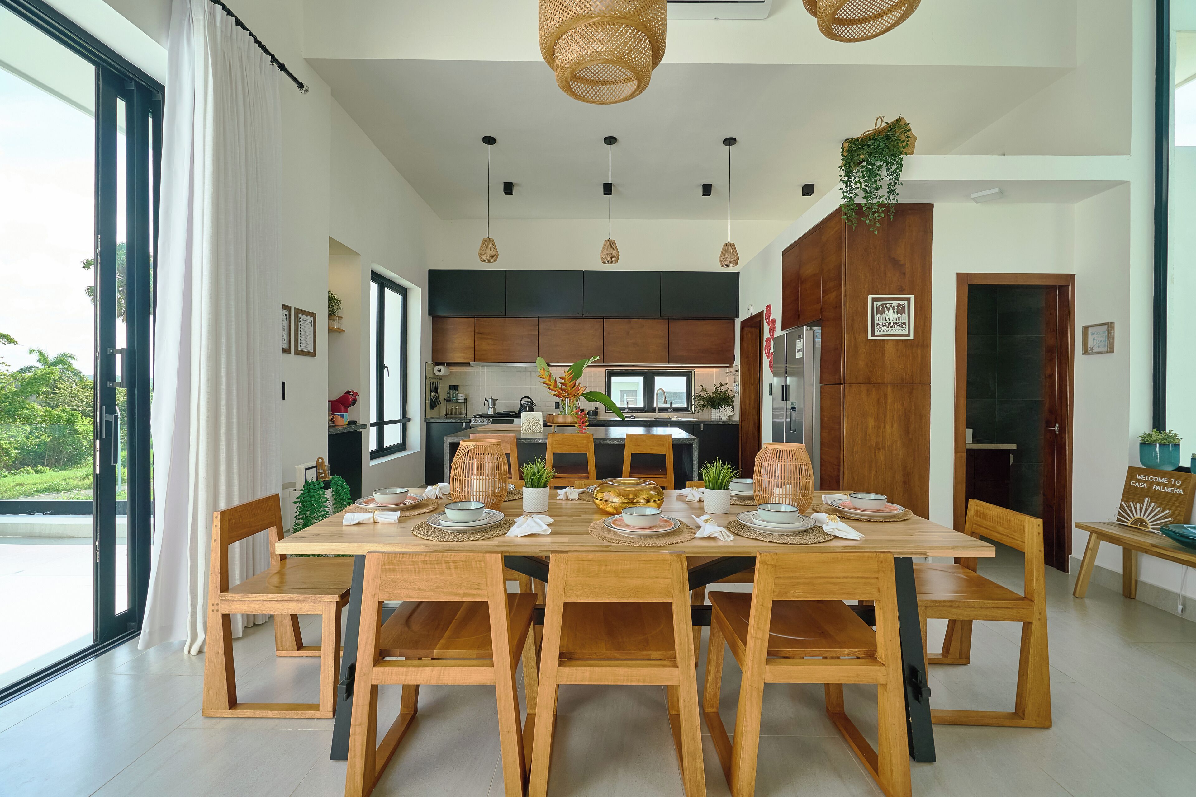 A dining area with a wooden table set for six, surrounded by chairs. There is a kitchen in the background with dark cabinets and a large window.