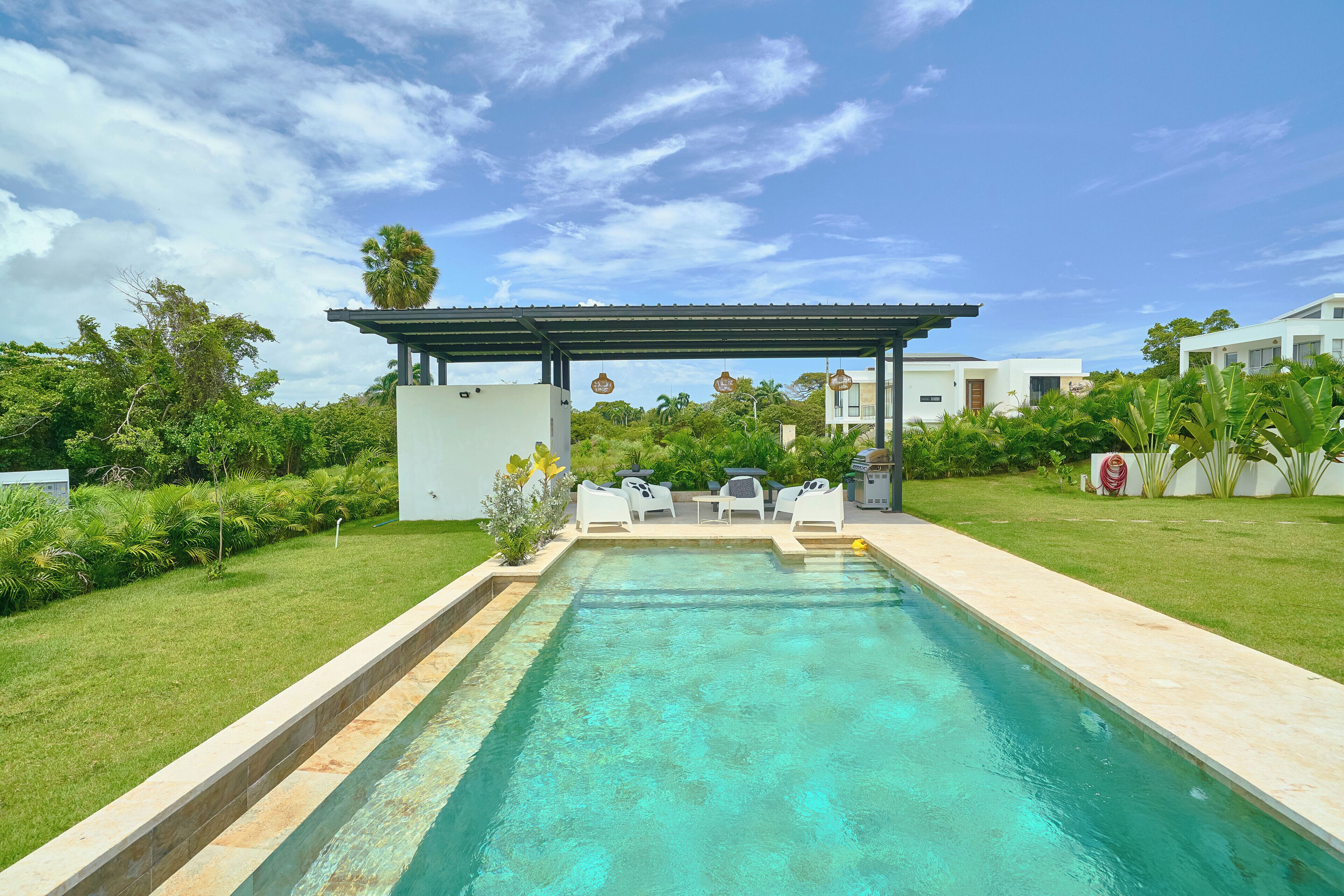 Ein Pool mit Pergola und Liegestühlen, umgeben von Grünflächen und modernen Gebäuden.