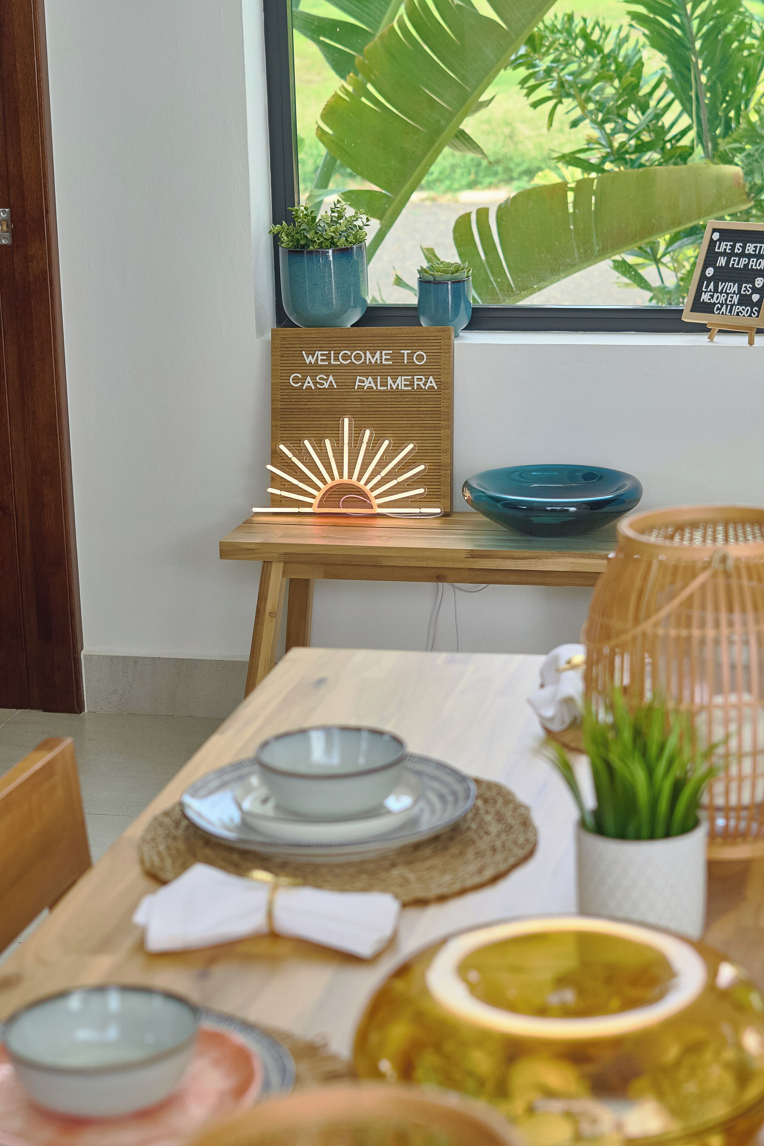 Un coin repas avec une table en bois dressée pour le repas, un panneau d’accueil et une vue sur la verdure à l’extérieur.