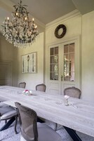 Un comedor con una larga mesa, sillas, una araña y una puerta con paneles de vidrio, además de una corona colgada en la pared.