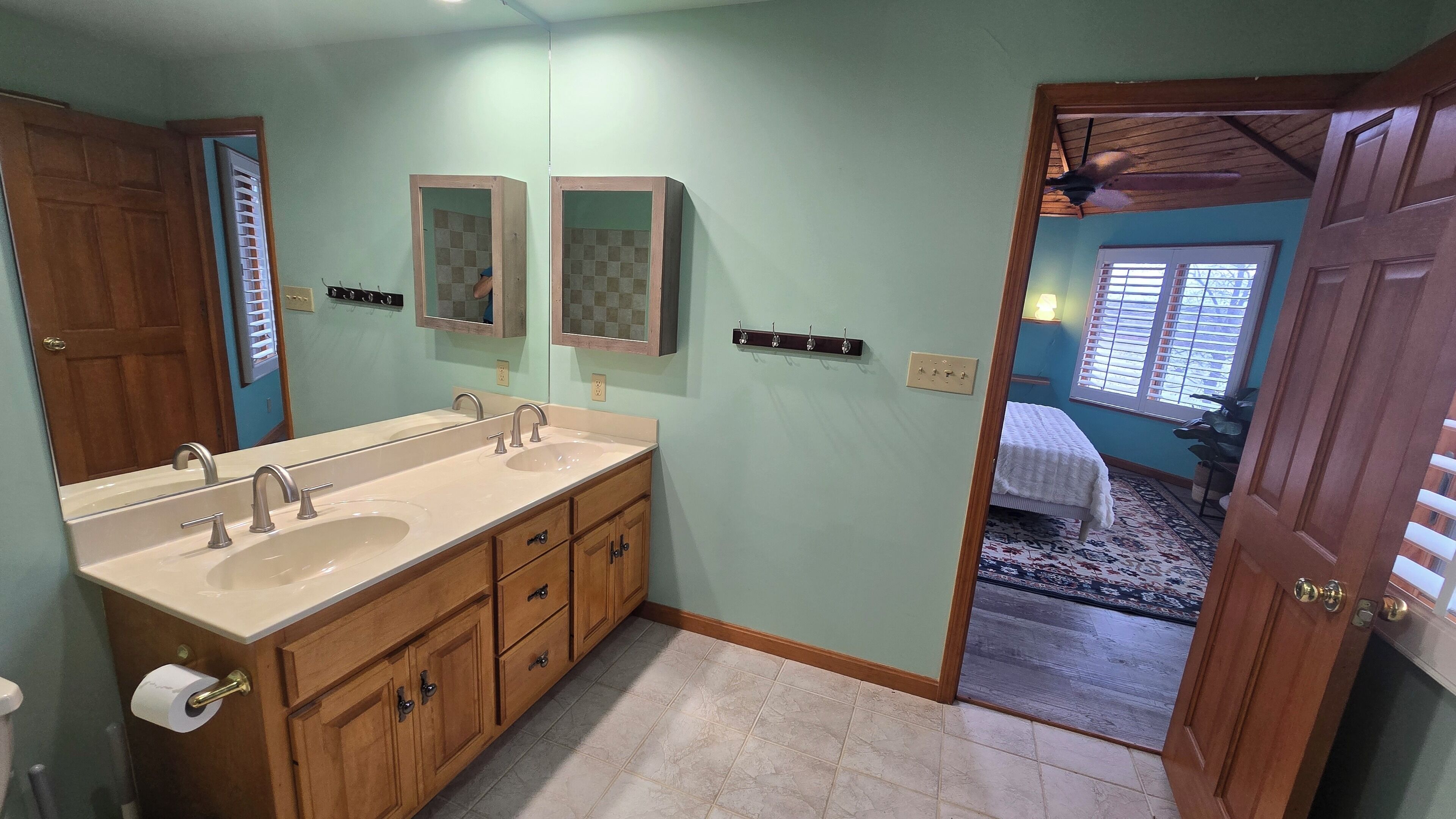 A bathroom with two sinks, wooden cabinets, a mirror, and a door leading to a bedroom.