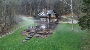 A two-story wooden house with a deck, surrounded by a grassy yard and a stone pathway.