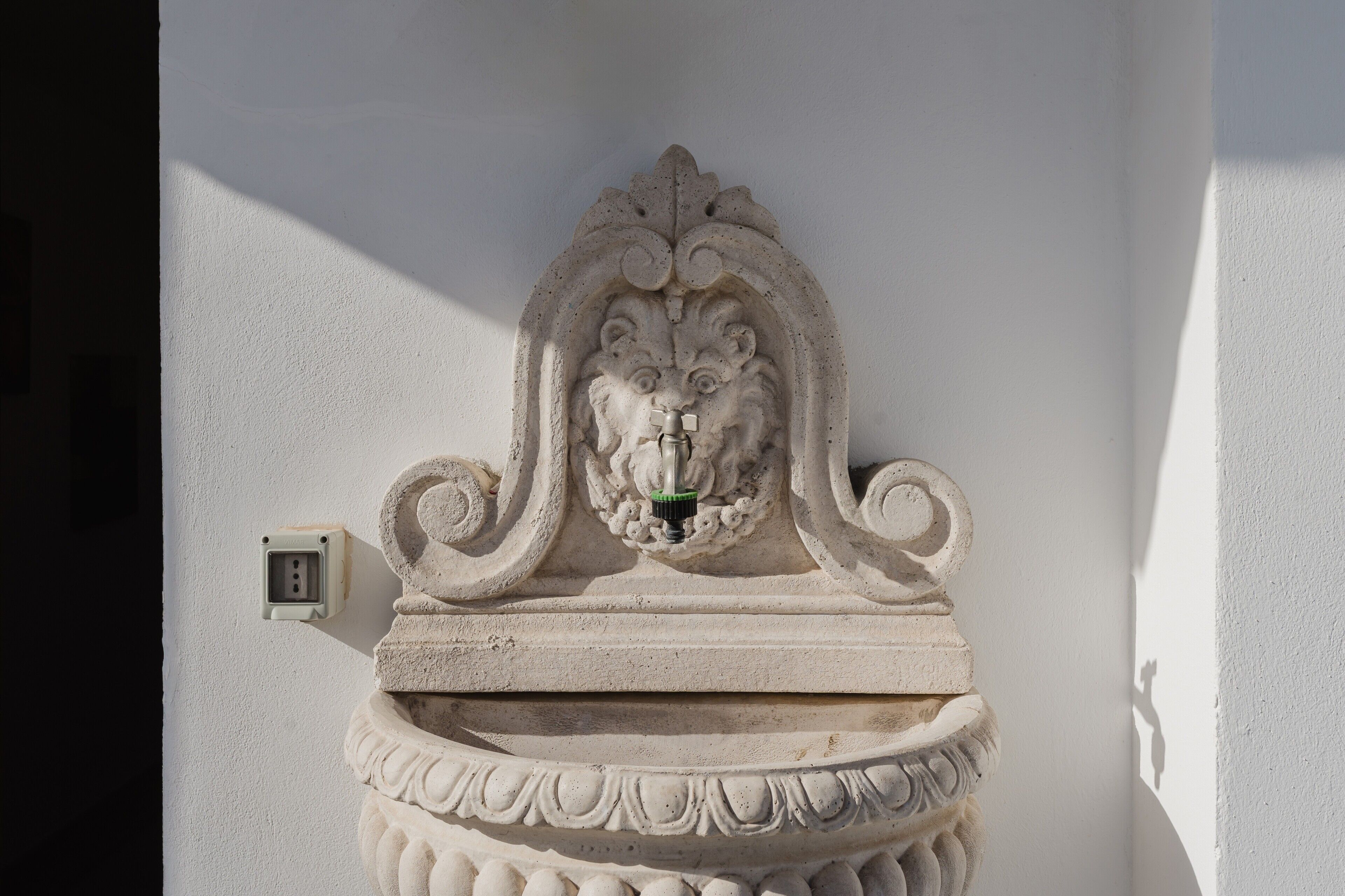 A stone wall-mounted water fountain with a lion's head design, a power outlet, and a textured wall.