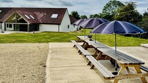 Ein Picknickplatz mit Holzbänken und Sonnenschirmen, ein Kiesweg und im Hintergrund ein Gebäude mit einem geneigten Dach.