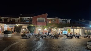 En brostensbelagt plads med udendørs siddepladser ved en restaurant, omgivet af bygninger med skilte.