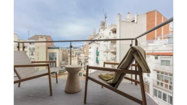 Un balcón con dos sillas de mimbre y una pequeña mesa redonda, con vistas a un paisaje urbano con edificios y cielo despejado.