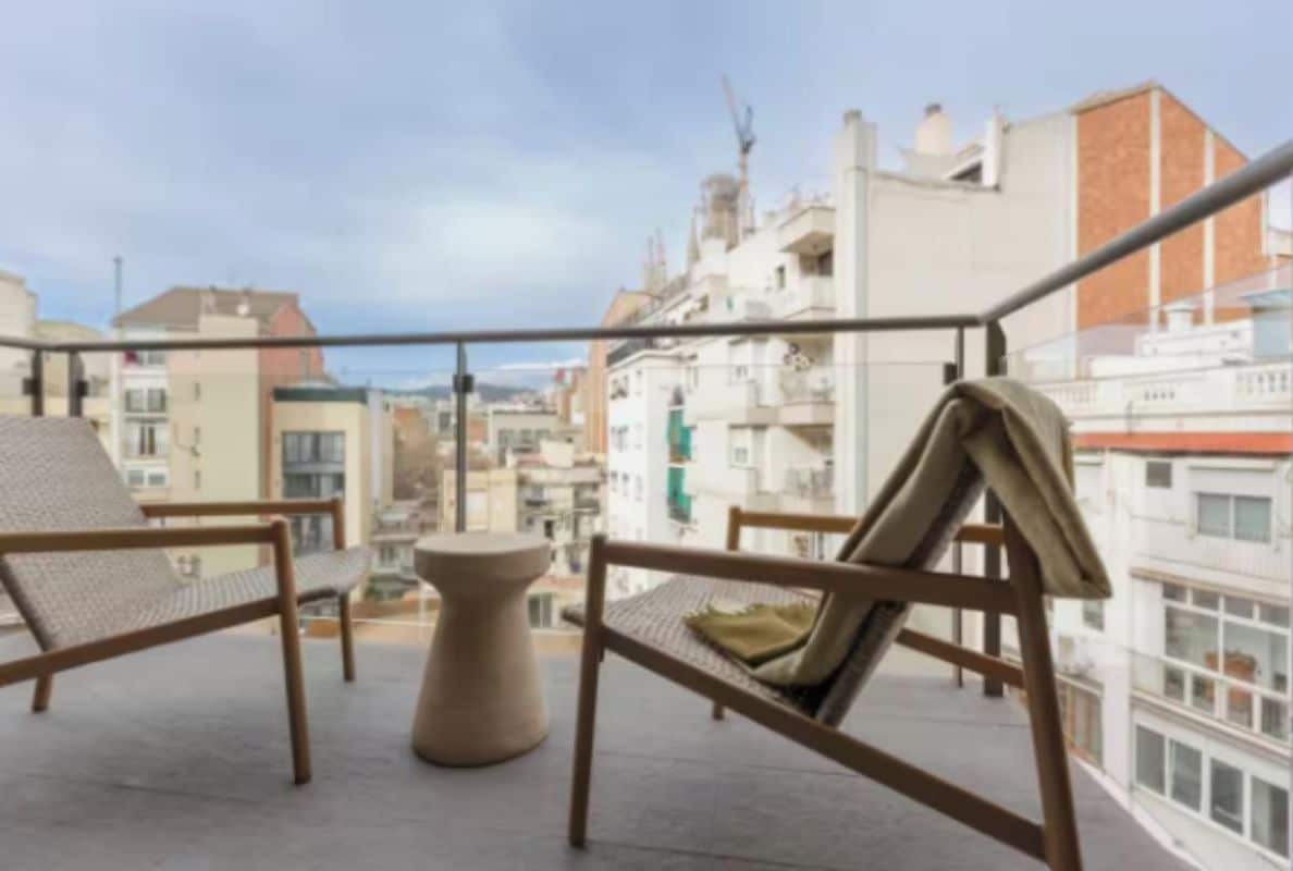 A balcony with two wicker chairs and a small round table, overlooking a cityscape with buildings and a clear sky.
