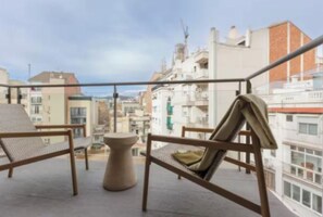 Un balcón con dos sillas de mimbre y una pequeña mesa redonda, con vistas a un paisaje urbano con edificios y cielo despejado.