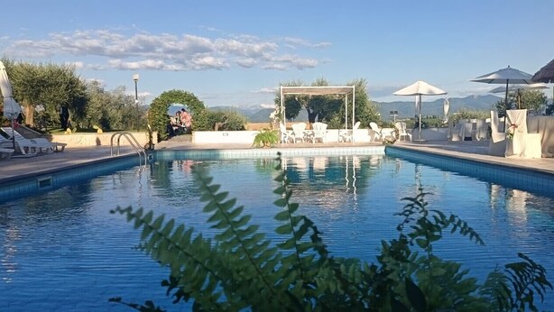 A swimming pool with lounge chairs and umbrellas, surrounded by greenery and a clear sky.