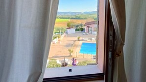 A window with curtains, a view of a swimming pool, and a building in the distance.