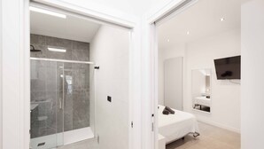 Un baño moderno con cabina de ducha de vidrio, una cama con colchón blanco y un televisor montado en la pared.