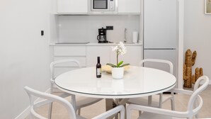 Una cocina moderna con gabinetes blancos, microondas, cafetera y refrigerador. Hay una mesa redonda de vidrio con sillas y una planta en maceta.