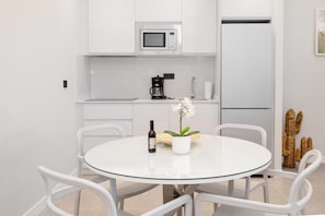 Eine moderne Küche mit weißen Schränken, einer Mikrowelle, einer Kaffeemaschine und einem Kühlschrank. Ein runder Glastisch mit Stühlen und einem Blumentopf steht ebenfalls dort.
