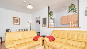 Ein Wohnzimmer mit gelbem Sofa, einem Glastisch, einer hängenden Pflanze und einer sichtbaren Küchenzeile im Hintergrund.