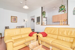 Un salon avec un canapé jaune, une table basse en verre, une plante suspendue et, au fond, un coin cuisine.