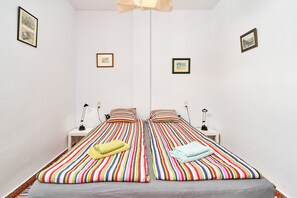 Una habitación con dos camas, cada una con ropa de cama y almohadas a rayas, una mesita con una lámpara y cuadros en las paredes.