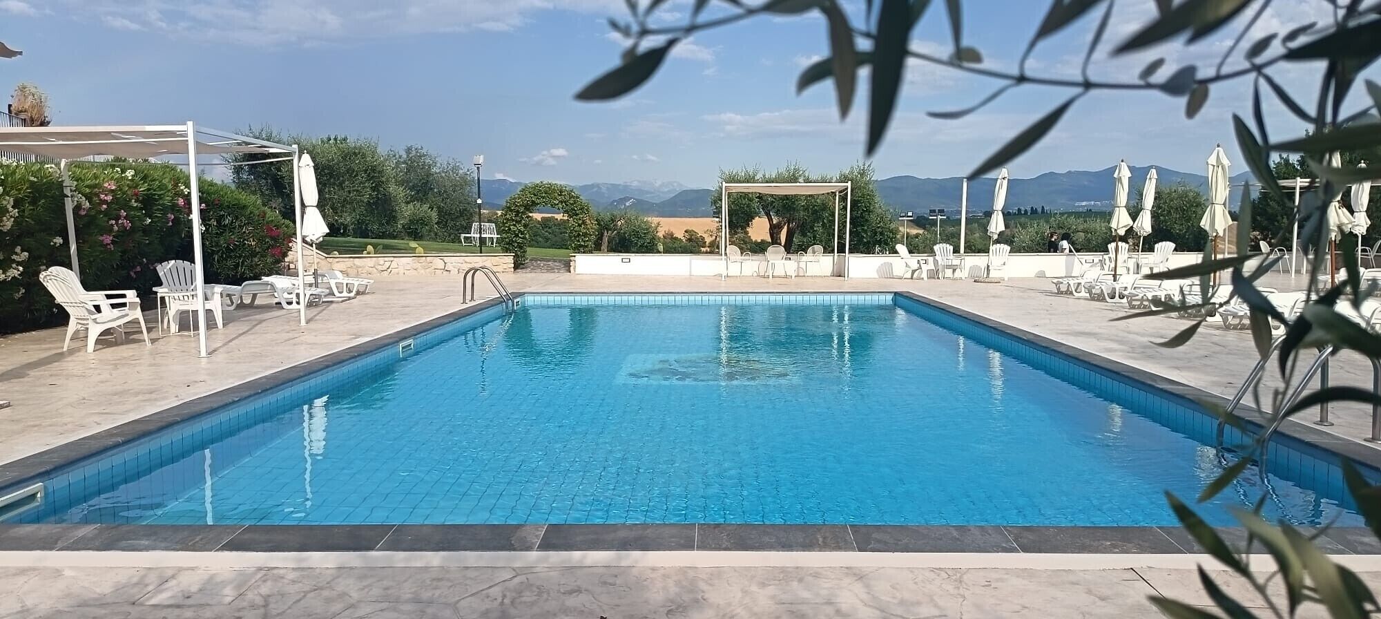A rectangular swimming pool with clear blue water, surrounded by white lounge chairs and umbrellas, set against a backdrop of mountains and clear sky.
