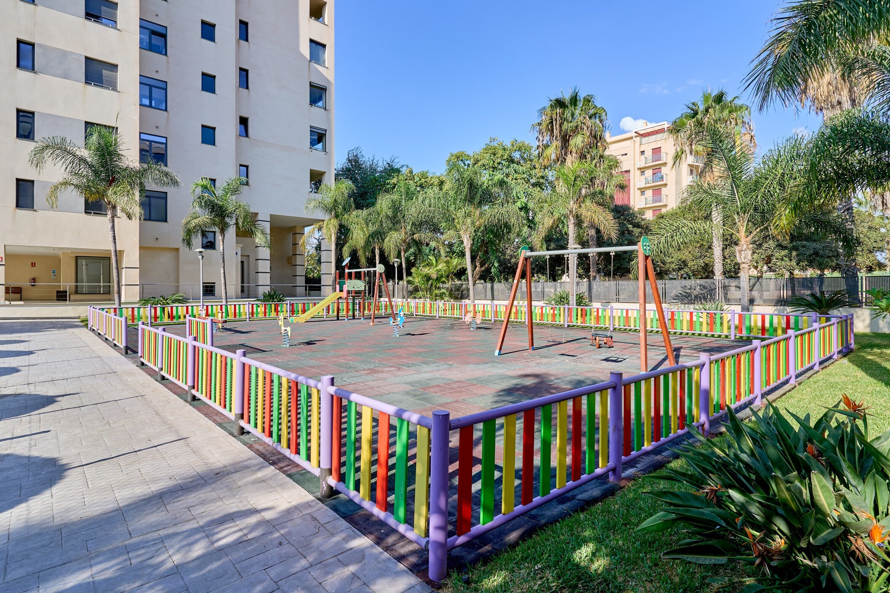 Un patio de recreo delimitado por una cerca colorida, rodeado de palmeras y un edificio de varios pisos.