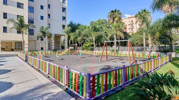 Un patio de recreo delimitado por una cerca colorida, rodeado de palmeras y un edificio de varios pisos.