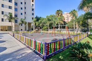 Un patio de recreo delimitado por una cerca colorida, rodeado de palmeras y un edificio de varios pisos.