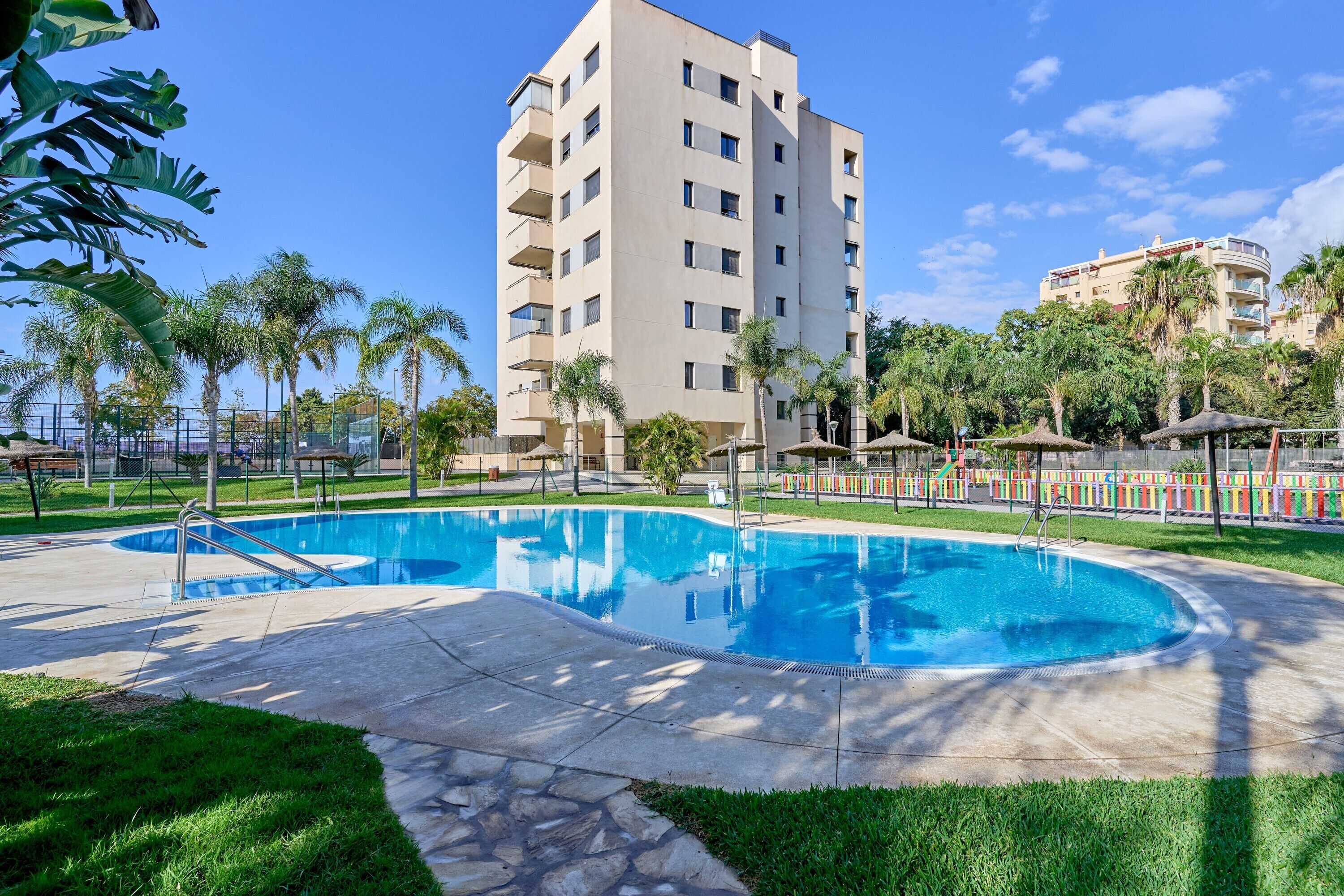 Una piscina rodeada de césped cuidado, palmeras y un edificio de varios pisos con balcones.