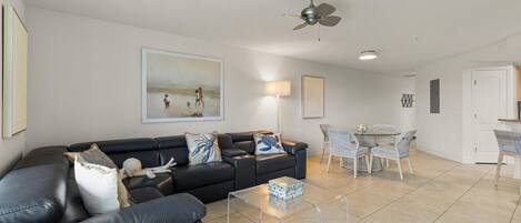 A modern living room with a black leather sofa, a glass coffee table, and a dining area with wicker chairs.