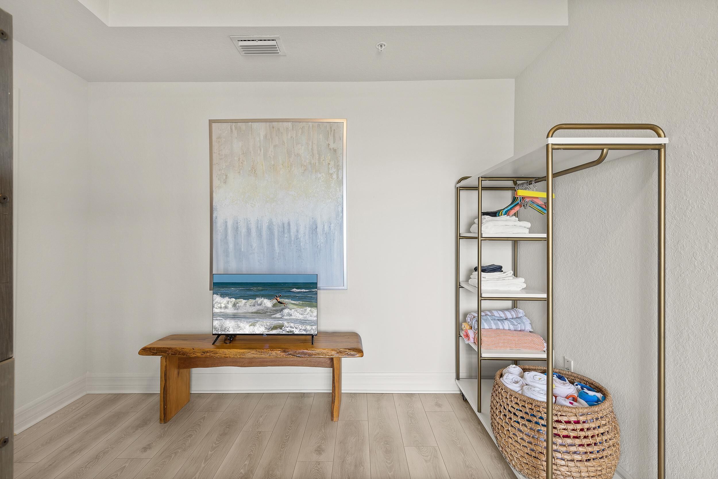 A room with a wooden bench, a large abstract painting, a metal shelving unit with folded clothes, and a woven basket.
