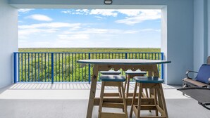 A balcony with a table and two chairs, overlooking a green landscape with a blue railing. - Oceanwalk 17-502 (New Smyrna Beach)