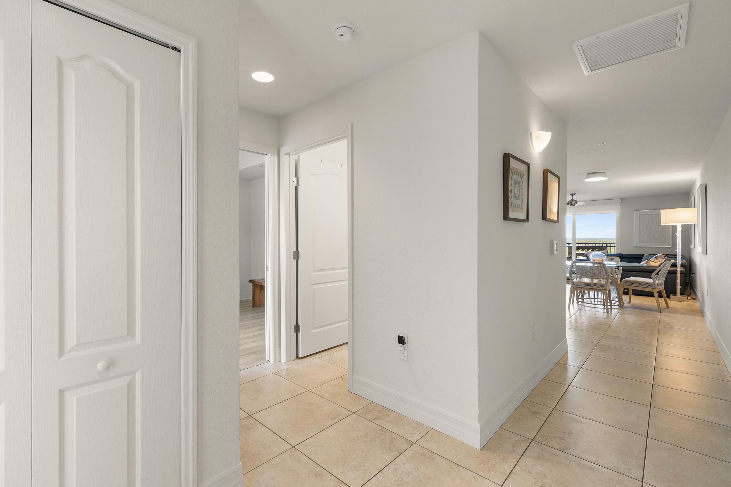 A hallway with white walls, a tiled floor, and a dining area with chairs and a table.