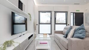 Una sala de estar moderna con un televisor de pantalla plana, una mesa de centro de vidrio, un sofá color beige con cojines y un ventanal con vistas a la ciudad.