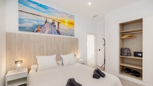 Una habitación de hotel moderna con una cama grande, una mesita de noche con lámpara, cabecera de madera, un cuadro de un atardecer y un vestidor con estantes y perchas.