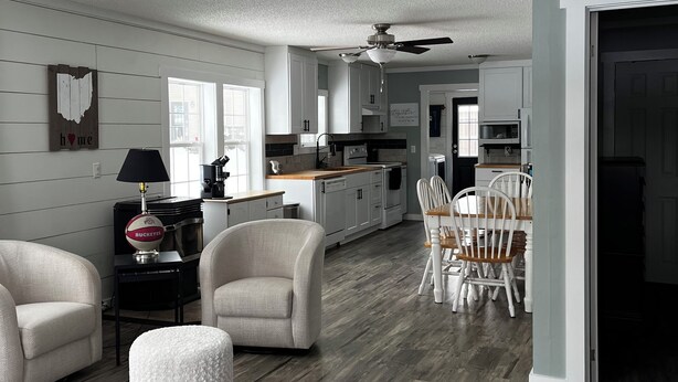 A modern kitchen with white cabinets, a dining area with a wooden table and chairs, and a living room with a sofa and ottoman.