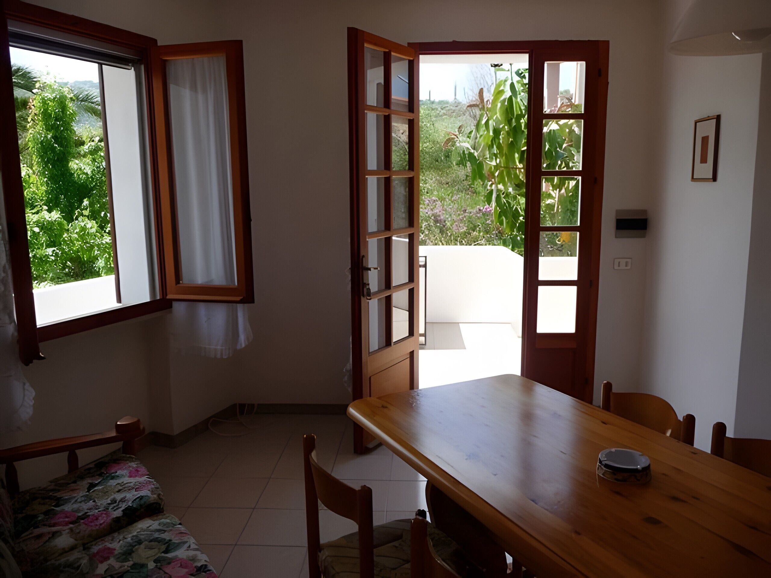 A room with a wooden dining table, a bench with a floral cushion, a window with curtains, and an open door leading outside.