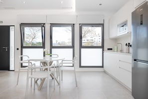 A modern kitchen with white cabinets, a stainless steel refrigerator, and a dining area with white chairs and a glass table.