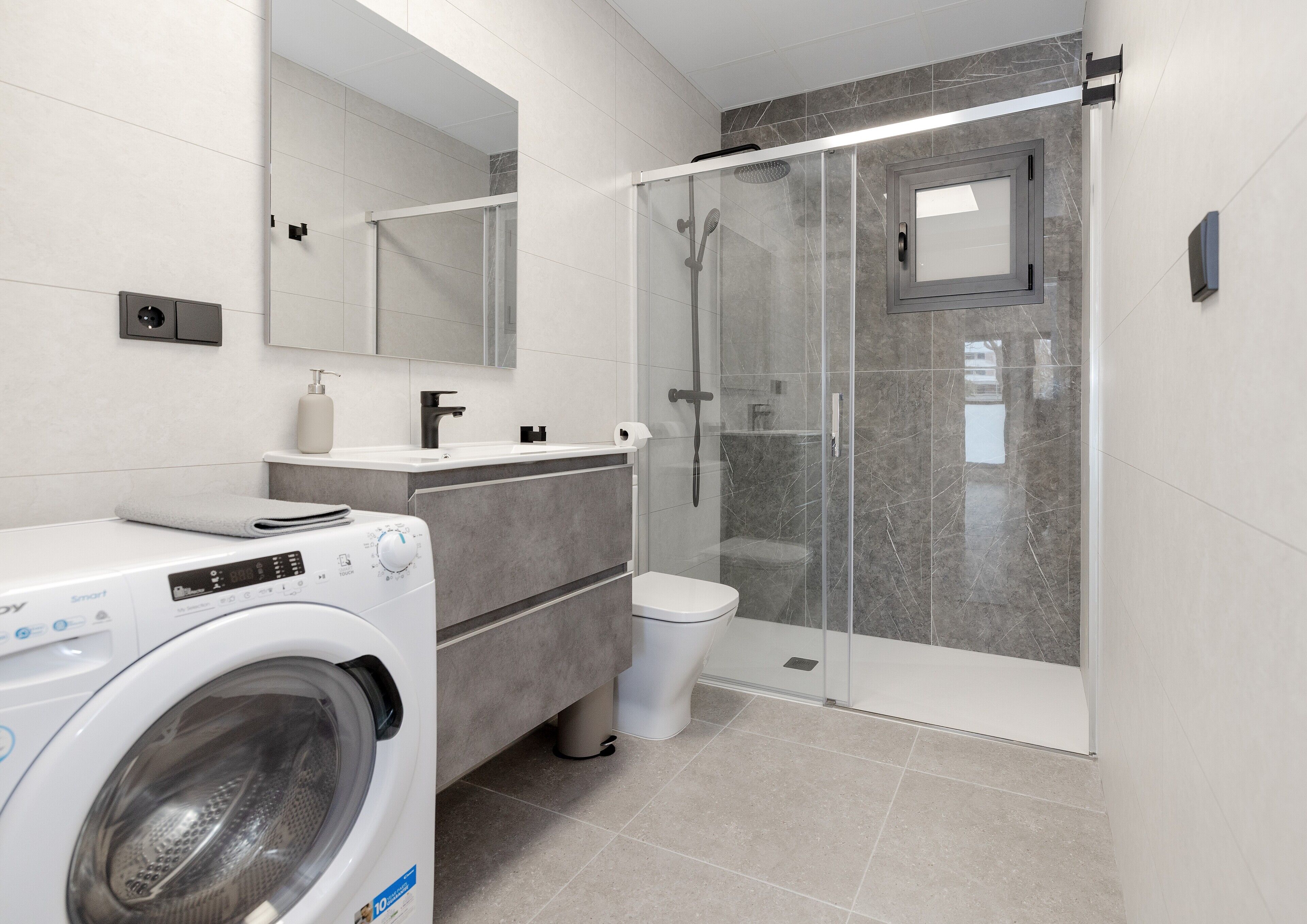 Une salle de bain moderne équipée d’un lave-linge, de toilettes, d’une douche à parois de verre et d’un espace de douche carrelé en marbre.