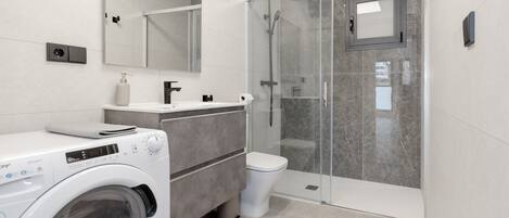 Une salle de bain moderne équipée d’un lave-linge, de toilettes, d’une douche à parois de verre et d’un espace de douche carrelé en marbre.