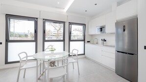 Une cuisine moderne avec une table en verre, des chaises blanches, un réfrigérateur en acier inoxydable et de grandes fenêtres.