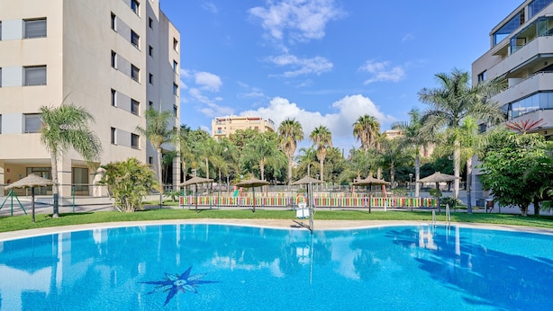 A swimming pool surrounded by palm trees and a multi-story building. - Apartment '2-Bed Antares' with Shared Pool, Wi-Fi, and Air Conditioning (Torre del Mar)